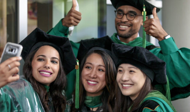 Four graduates dressed in regalia smile into a cell phone for a selfie