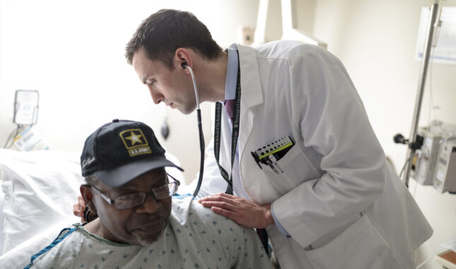 An M4 student in a white coat places one had on a patient's shoulder and uses the other to listen to his breathing