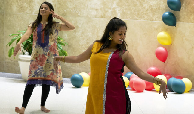 Two students in brightly colored dresses smile and dance for a performance